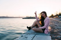 Улыбающаяся молодая женщина, сидящая на набережной рядом с поверхностью воды с кораблем на закате и показывающая знак ОК — стоковое фото