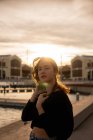 Прекрасна молода жінка дивиться на камеру, стоячи біля води на міській набережній під час заходу сонця — стокове фото