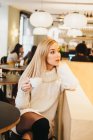 Attraktive, leidenschaftliche Dame im Strickkleid hält Becher mit Heißgetränk in der Hand und schaut im Café weg — Stockfoto