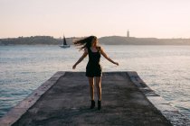 Donna allegra in abito nero e stivali che ballano su argine vicino alla superficie dell'acqua con yacht al tramonto — Foto stock