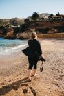 Visão traseira da jovem senhora descalça com câmera de fotos andando na areia perto de água do mar — Fotografia de Stock