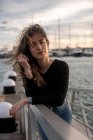 Porträt einer jungen Frau, die lockiges Haar berührt und in die Kamera blickt, während sie sich an ein Geländer auf verschwommenem Hintergrund aus Seebrücke und Wasser lehnt — Stockfoto