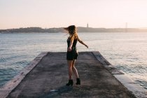 Cheerful woman in black wear and boots dancing on embankment near water surface at sunset — Stock Photo