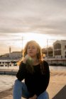 Прекрасная молодая женщина, поддерживающая голову и смотрящая в камеру, сидя возле воды на городской набережной во время заката — стоковое фото