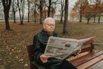 Hombre mayor en chaqueta de cuero leyendo periódico fresco mientras está sentado en el banco en el parque de otoño - foto de stock