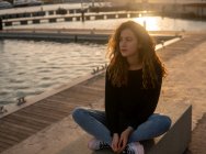 Jovem pensativa olhando para a câmera enquanto se senta perto da água no dique da cidade durante o pôr do sol — Fotografia de Stock