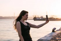 Jolie jeune femme en robe noire posant et prenant selfie sur le remblai près de la surface de l'eau au coucher du soleil — Photo de stock