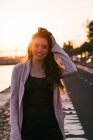 Портрет улыбающейся привлекательной молодой женщины с рукой в волосах, стоящей на набережной возле поверхности воды на закате — стоковое фото
