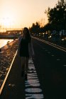 Silhouette de jeune fille en robe et bottes marchant sur l'allée près de l'eau au coucher du soleil — Photo de stock