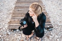 From above shot of pretty female with photo camera smiling and looking away while sitting on rocky coast in Altea, Spain — Stock Photo