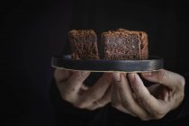 Mani umane in possesso di un piatto con pezzi di cioccolato brownie su sfondo scuro — Foto stock
