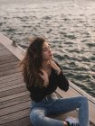Affascinante giovane femmina con gli occhi chiusi seduta su un molo di legno vicino all'acqua — Foto stock