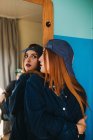 Jeune femme charmante aux cheveux rouges en tenue décontractée avec la main croisée regardant le reflet près du mur bleu — Photo de stock