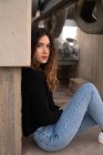 Чарівна молода жінка з кучерявим волоссям дивиться на камеру, сидячи біля стіни бетонної будівлі — стокове фото