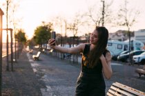 Attractive lady in black dress with hand in hair taking selfie on street at sunset — Stock Photo