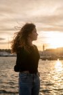 Attrayant jeune femme avec les cheveux bouclés regardant loin tout en se tenant près de l'eau pendant le coucher du soleil en ville — Photo de stock