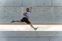 Attractive bearded man in sportswear smiling and jumping high during outdoor training on street — Stock Photo