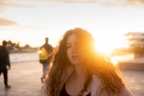 Jeune femme avec les cheveux bouclés regardant la caméra tout en se tenant sur la rue de la ville pendant le coucher du soleil — Photo de stock