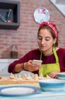 Adolescente utilisant le smartphone tout en travaillant sur la pâtisserie traditionnelle — Photo de stock