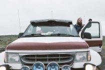 Hombre barbudo adulto de pie cerca del coche en la carretera fría remota mientras viaja, Islandia - foto de stock