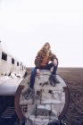 Jeune femme en tenue chaude assise sur un aéronef brisé entre des terrains sombres en Islande — Photo de stock
