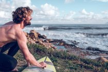 Young man cleaning surfboard on ocean coast and looking at view — Stock Photo