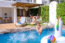 Garoto bonito em shorts de natação e óculos pulando na água da piscina enquanto passa tempo com a mãe no resort — Fotografia de Stock
