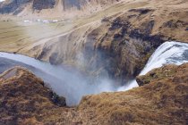 Над водой каскад падает в горной реке между коричневыми каменными холмами в Исландии — стоковое фото