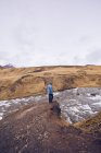 Парень с боковым видом, стоящий на камне возле течения реки между коричневыми горами в Исландии — стоковое фото