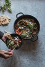 Human hands holding bowl over pot of ragout with lentil and sweet potato curry and bowl on grey tabletop — Stock Photo