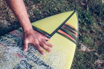 Close-up of male hand on colorful surfboard with crumbs of wax on grass — Stock Photo