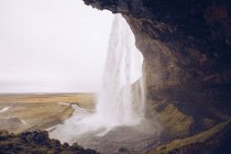 Caverna com cascata de água caindo no rio que corre entre terras selvagens na Islândia — Fotografia de Stock