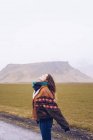 Vista posterior joven atractiva dama en? coat mirando la cámara en el camino entre tierras salvajes con colinas de piedra en Islandia - foto de stock