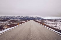 Деревенская дорога между дикими землями в снегу, ведущая к горам и небу в облаках Исландии — стоковое фото
