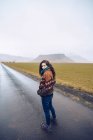 Vue de dos jeune femme attrayante en manteau regardant la caméra sur la route entre les terres sauvages avec des collines de pierre en Islande — Photo de stock