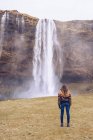 Voltar vista jovem senhora no campo perto de cascata de água caindo no rio entre rochas na Islândia — Fotografia de Stock