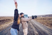 Vista laterale signora sorridente con mano alzata vicino ragazzo prendere selfie sulla macchina fotografica vicino auto sulla strada tra terre selvagge in Islanda — Foto stock