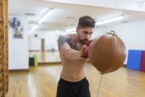 Jeune homme torse nu boxe avec sac de boxe dans la salle de gym — Photo de stock