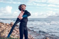 Молодий бородатий чоловік одягає гідрокостюм біля океану — стокове фото