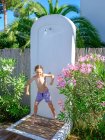 Mignon garçon en short de bain souriant et debout sous l'eau de douche par une journée ensoleillée dans une merveilleuse station balnéaire — Photo de stock