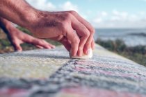 Primer plano de las manos masculinas extendiendo cera en la tabla de surf en la hierba cerca del mar - foto de stock