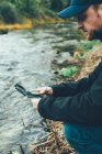 Giovane uomo che tiene una bussola in montagna accanto a un fiume con sfondo di colore marrone — Foto stock