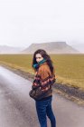Visão traseira jovem senhora atraente olhando para a câmera na estrada entre terras selvagens com colinas de pedra na Islândia — Fotografia de Stock
