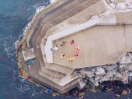 From above shot of anonymous people forming circle while standing on concrete building on amazing rocky cliff near waving sea — Stock Photo