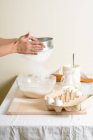 Mains de femme méconnaissable tamiser la farine sur un bol avec de la pâte dans une cuisine confortable . — Photo de stock