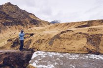 Vista lateral cara de pé na pedra perto de rio de streaming entre montanhas marrons na Islândia — Fotografia de Stock