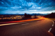 Вид на яскраві сліди світла на маршруті ввечері в сільській місцевості на фоні міста і хмарне небо — стокове фото