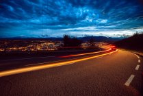 View of bright trail lights on route in evening in countryside on city background and cloudy sky — Stock Photo