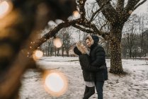 Side view of happy guy hugging young lady in ski jackets near wood decorated by illuminated fairy lights — Stock Photo