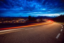 Vue des lumières du sentier lumineux sur la route en soirée à la campagne sur fond de ville et ciel nuageux — Photo de stock